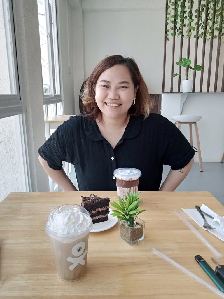 An image of a woman sitting in a black top in a cozy coffee shop, with a table featuring delicious coffee and cake