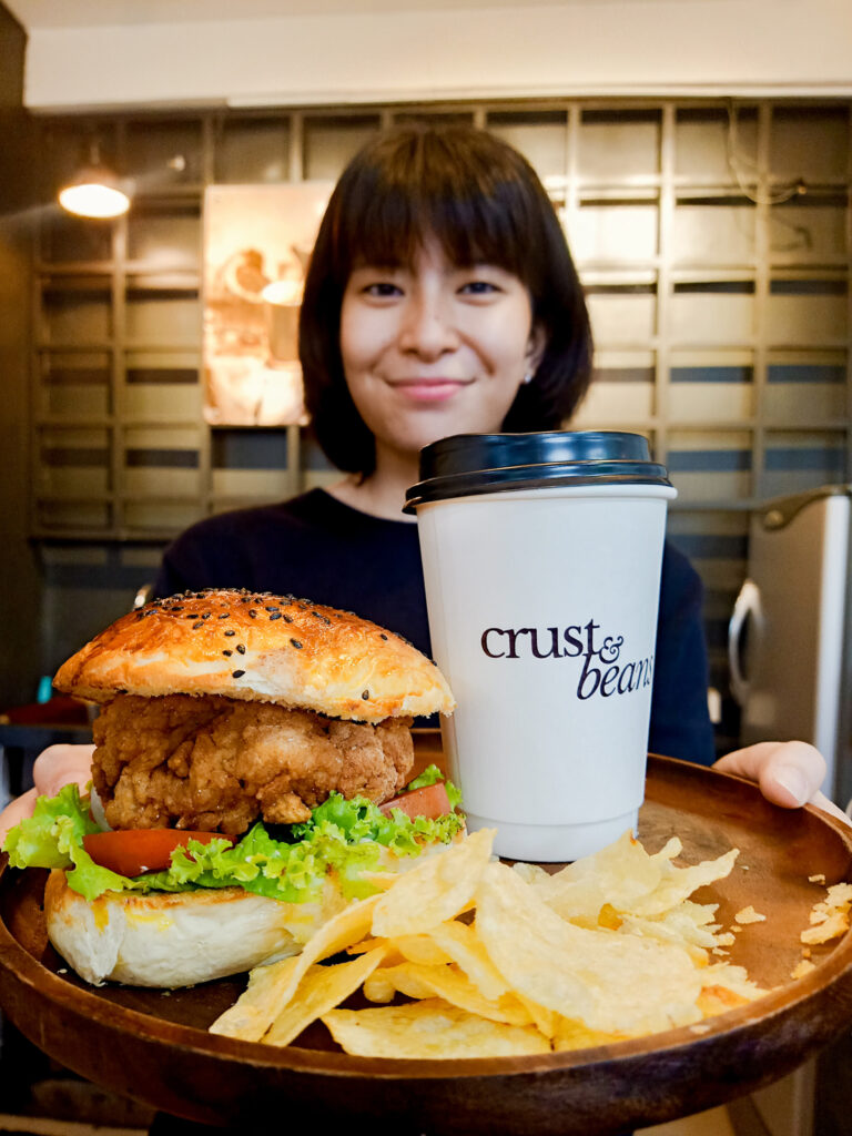 An image of crust and beans coffee with chicken burger on the tray. Serving by a female crew in black top