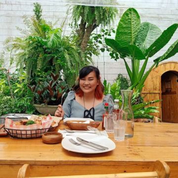 A woman sits at a table in an outdoor coffee shop nearby, surrounded by lush green plants. She smiles as she is about to enjoys her delicious meal. The sun shines down on the scene, casting a warm glow on the woman's face and highlighting the vibrant colors of the plants in the background.